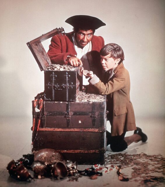 Robert Newton in a red coat and a pirate hat, and young Bobby Driscoll, looking into a box of pirate treasure. This story includes an early example of the phrase "shiver me timbers"