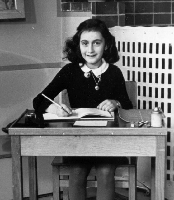 Anne Frank sitting behind a desk holding a pen, smiling.