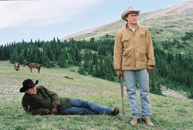 Jake Gyllenhaal lying on a grassy hill while Heath Ledger stands beside him holding a gun. Both men wear cowboy hats. 