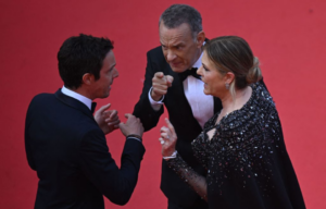 Tom Hanks aggressively points at a man in a suit while Rita Wilson talks to him on the red carpet.