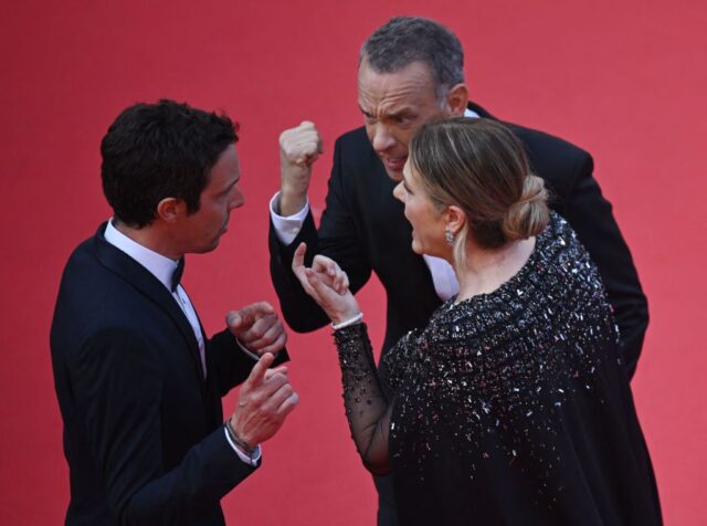 Tom Hanks and Rita Wilson appear to be yelling and gesturing at a man in a suit on the red carpet.