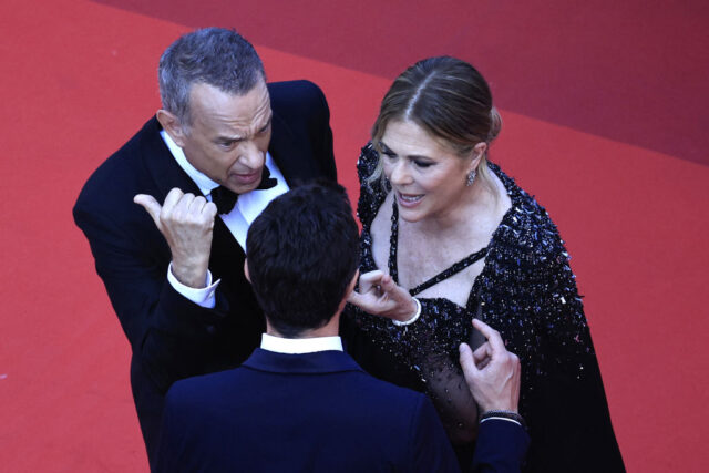 Tom Hanks and Rita Wilson appear to be yelling and gesturing at a man in a suit on the red carpet.