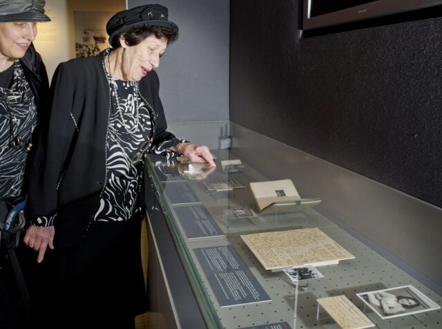 Hannah Pick-Goslar in a black hat and black clothing pointing at something in a glass case.
