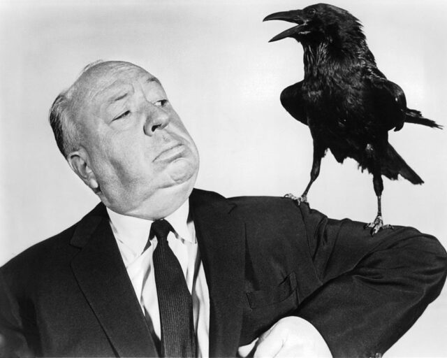 Alfred Hitchcock in a black suit and tie lifts his arm where a stuffed crow sits. 