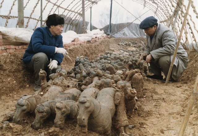 Two men crouch down beside the tops of small terracotta figures sticking out of the ground.