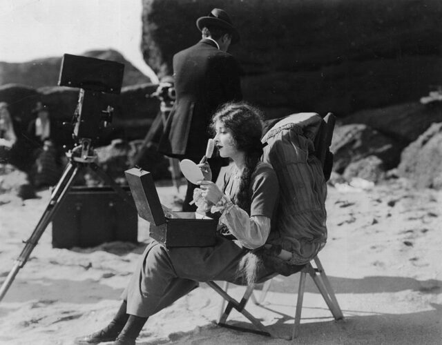 Mildred Harris applying makeup while sitting in a chair on the beach