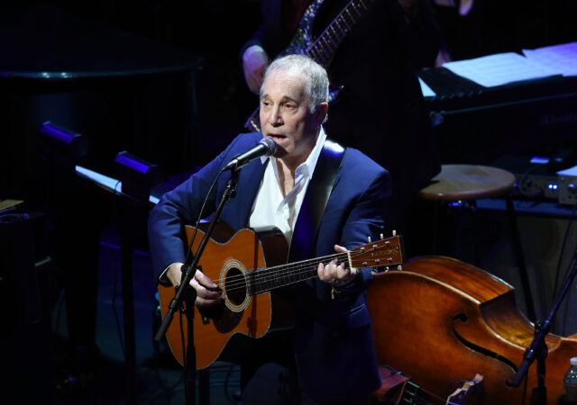 Paul Simon playing the guitar and performing on stage.