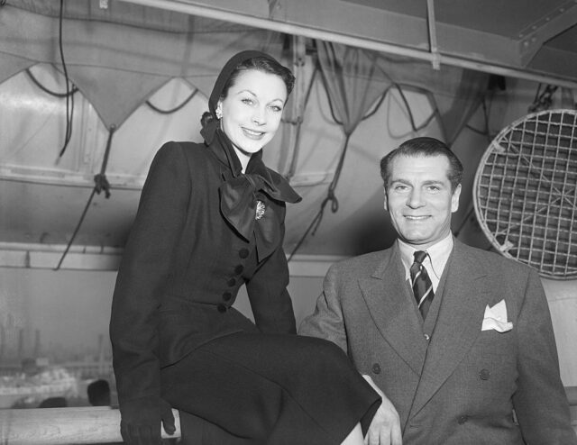 Vivien Leigh in all black sits on a railing beside Laurence Olivier who wears a grey suit.