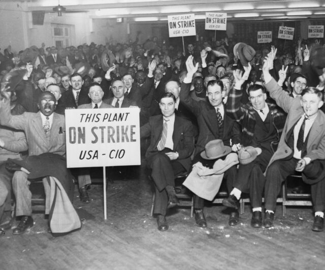 Workers sitting in a room, several strike signs being held.
