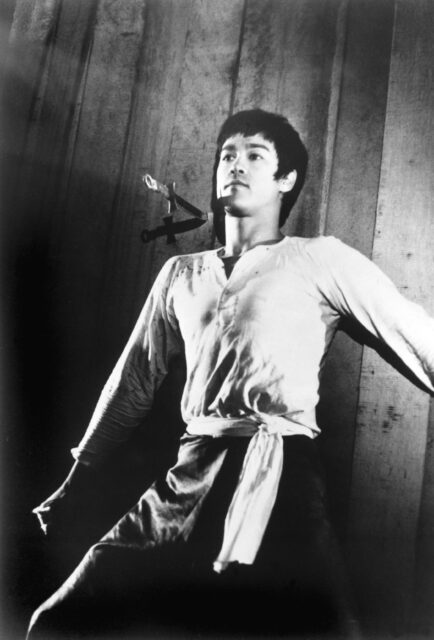 Bruce Lee laying on the floor, a dagger in the floor beside his head