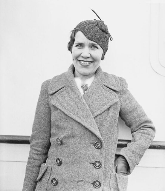 Maria Rasputin in a button-up jacket and hat, leaning against a railing while she smiles.