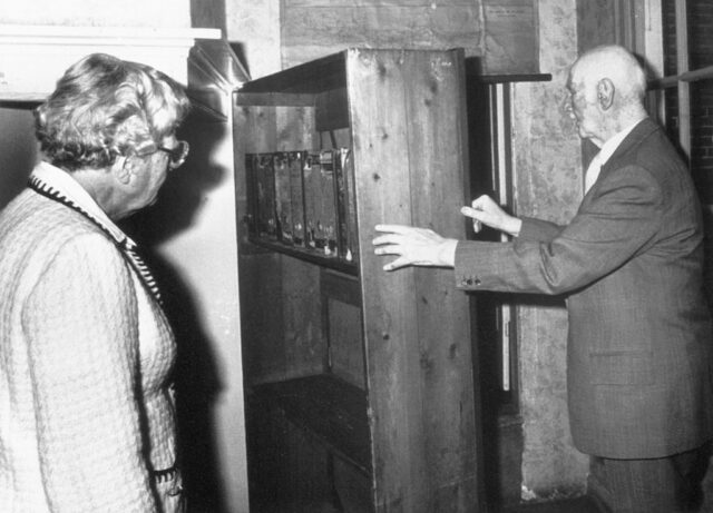 Otto Frank in a suit opens the wooden bookcase door that leads to the Secret Annex while Queen Juliana watches.