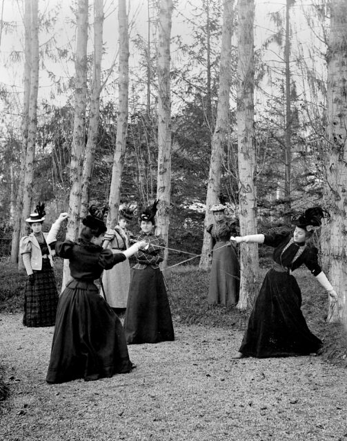 A group of Victorian women gathered in a forest, two are sword fighting.