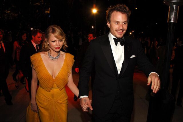 Michelle Williams in a yellow dress holding hands with a smiling Heath Ledger in a black suit as they walk the red carpet. 