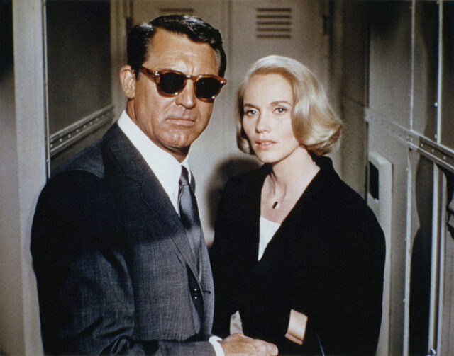 Cary Grant in a suit and black sunglasses stands with Eva Marie Saint in a black jacket.