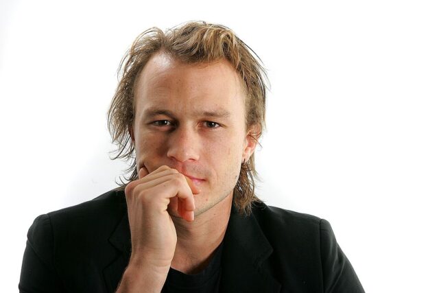 Heath Ledger resting his chin on his hand while wearing a black suit.