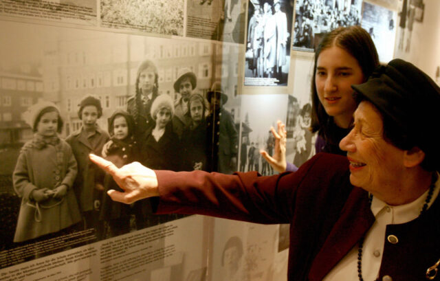 Hannah Pick-Goslar in a red jacket and black hat, points to a child in a large black and white photo on a wall.