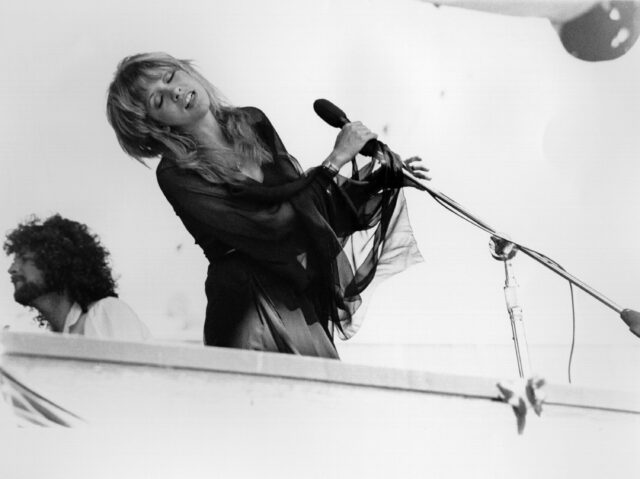 Stevie Nicks leaning back from a microphone, Lindsey Buckingham in the background.