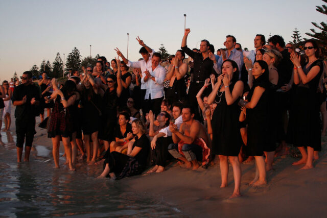 Large group of people wearing black sit and stand on a beach to watch the sunset.