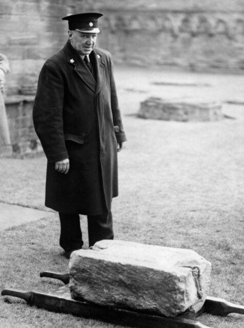 A police officer stands over the Stone of Destiny on a gurney on the ground.