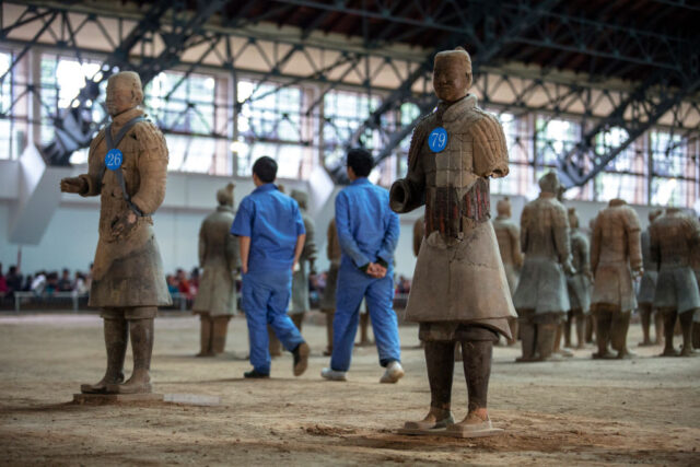 Two archaeologists in all blue walk through Terracotta warriors with number tags on their chests. 