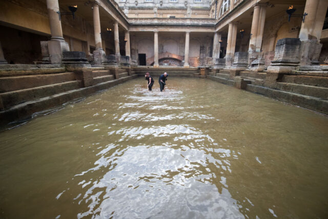 Two people in jumpsuits stand inside a Roman bath while they scrub the stone. 
