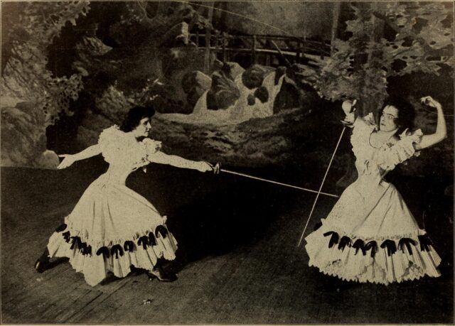 Two women in tight dresses perform a sword fight on stage.