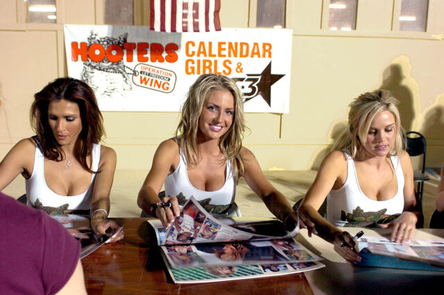 Three Hooters Girls sit at a table signing autographs