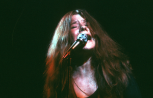 Headshot of Janis Joplin singing into a microphone, her hair slightly covering her face.