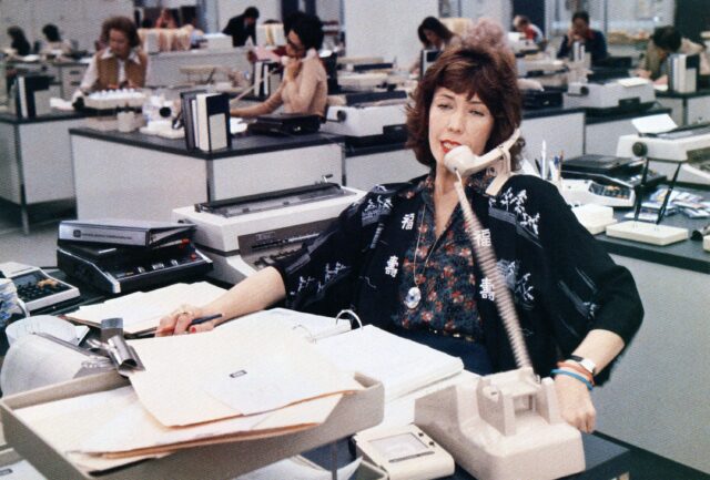 Lily Tomlin sitting at a desk covered in papers, speaking on the phone