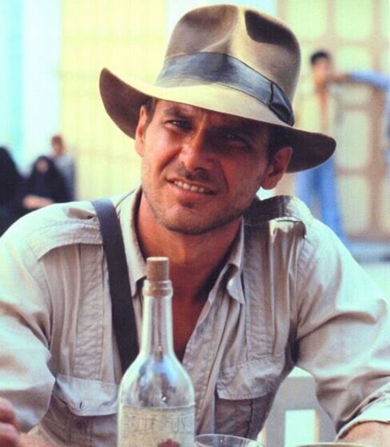 Harrison Ford dressed as Indiana Jones, a bottle in front of him