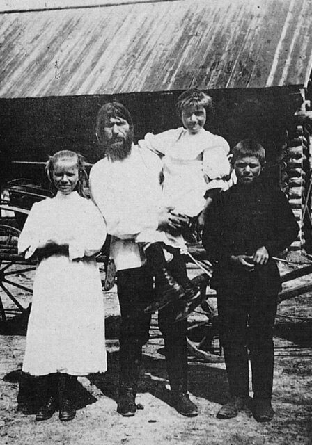 Grigori Rasputin in a white shirt and black pants holding a daughter, while a daughter and son stand with him in front of a wooden fence.