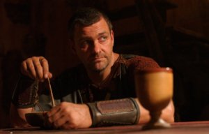 Ray Stevenson in Rome costume, sitting at a table.