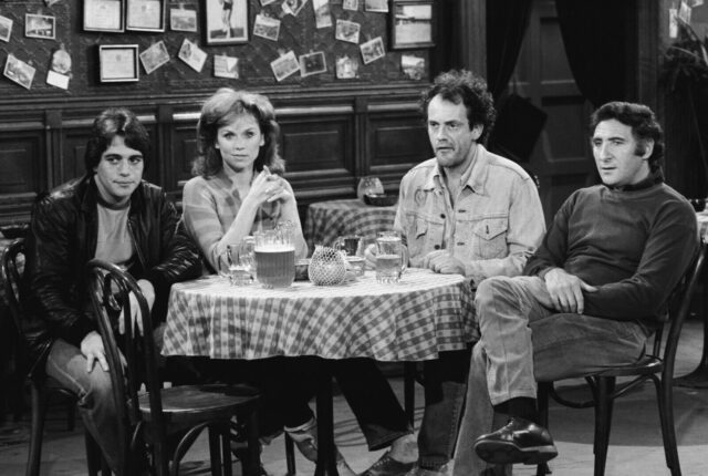The cast sitting at a table in a black and white promotional shot