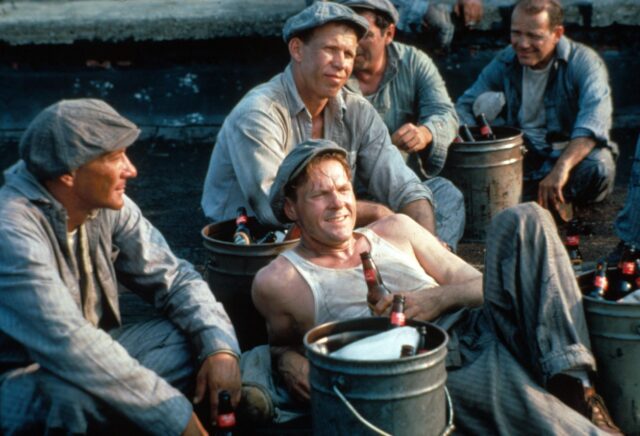 Several men sitting around drinking beers in 1950s prison attire.