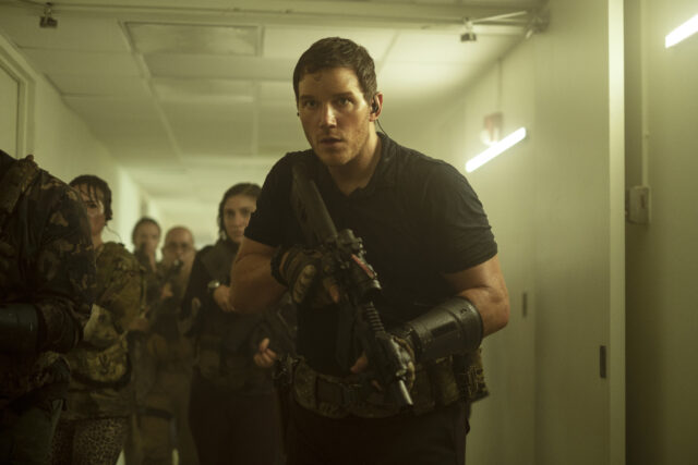 Chris Pratt in a hallway, holding a weapon, people walking behind him