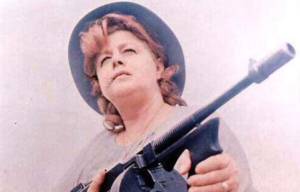 Shelley Winters as Ma Barker in a hat, holding out a gun.