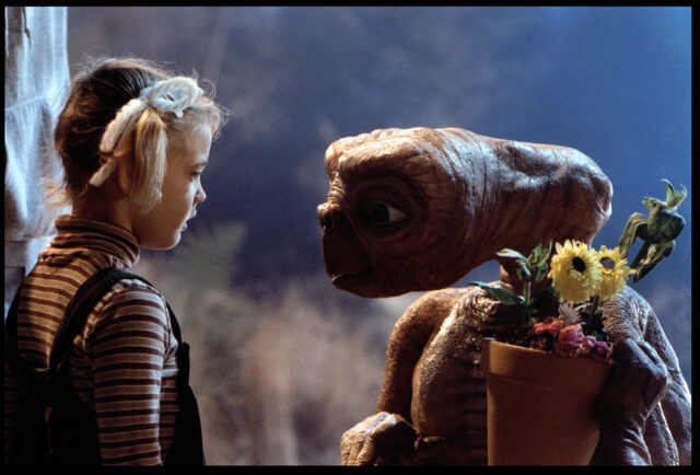 Drew Barrymore looking at E.T. holding a potted plant.