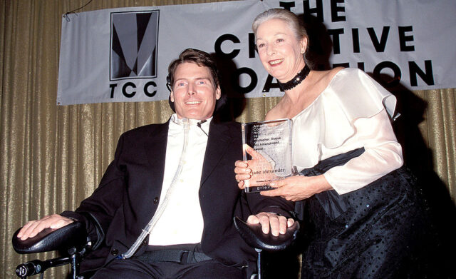 Christopher Reeve sitting in a wheelchair wearing a black suit beside a woman in a black and white dress holding up an award. 