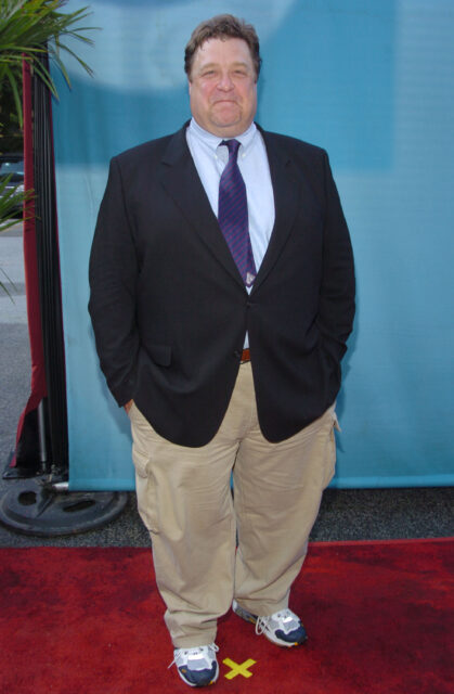 John Goodman standing with his hands in his pockets on a red carpet.