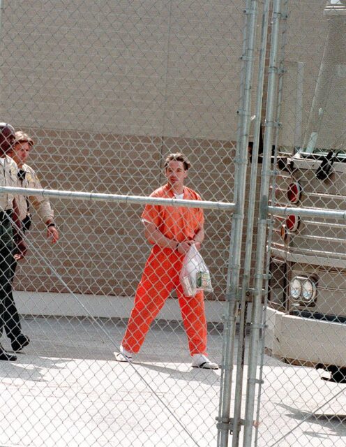 Young Robert Downey Jr. wearing an orange prison uniform and holding a bag as he walks towards a bus.
