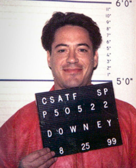 Young Robert Downey Jr. posing for a mug shot in an orange jumpsuit. 