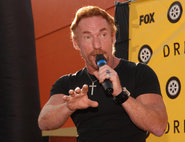 Danny Bonaduce speaking into a microphone and holding his hand forward.