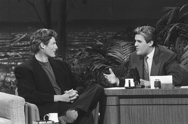 Christopher Reeve in a black jacket seated looking at Jay Leno who sits behind a desk wearing a jacket.