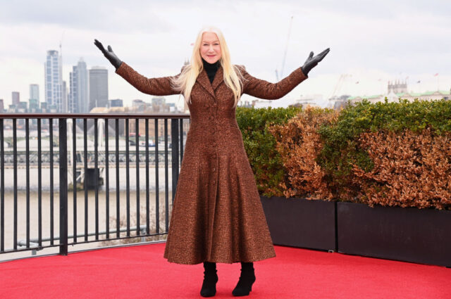Helen Mirren holding out her arms while wearing a long brown jacket.