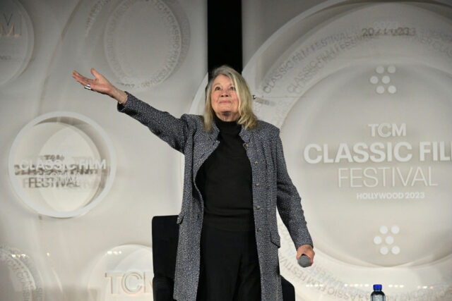 Angie Dickinson looking up to the sky and holding out one arm, wearing a black and white jacket and black shirt and pants.