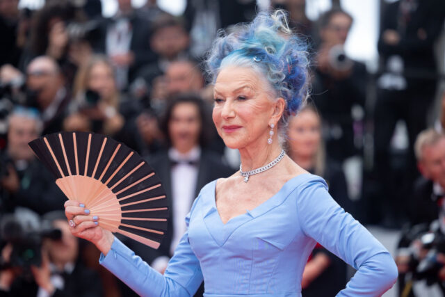 Helen Mirren holding a fan in a blue dress with her hair dyed a matching color of blue.