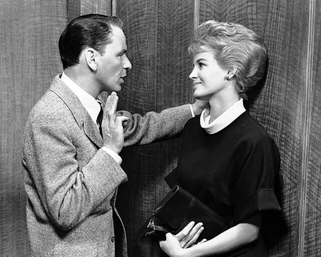 Frank Sinatra in a tweed jacket rests his arm beside Angie Dickinson's head. She looks at him while wearing a black dress and holding a purse.