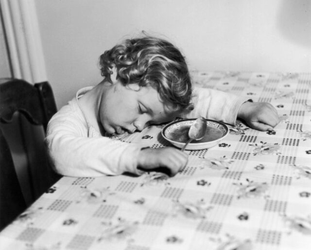 Young girl asleep at a table holding her spoon in front of a bowl of soup.