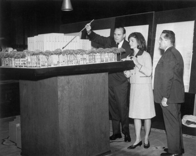 Jackie Kennedy, Jack Warnecke, and another man stand around a model of a building. Warnecke using a stick as a pointer.